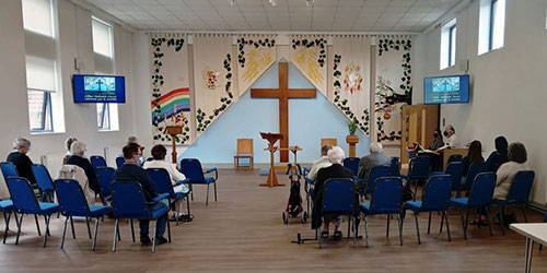 Church hall showing Methodist congregation sitting on plastic chairs. A cross is visible at the front